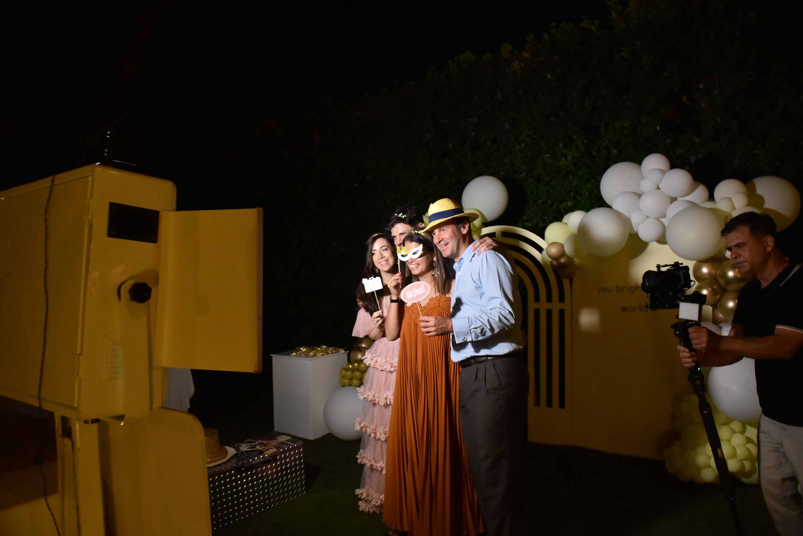 Four people posing with photo booth props including a white mask and an 'Aloha!' sign at a party with white and gold balloons, while a cameraman records.