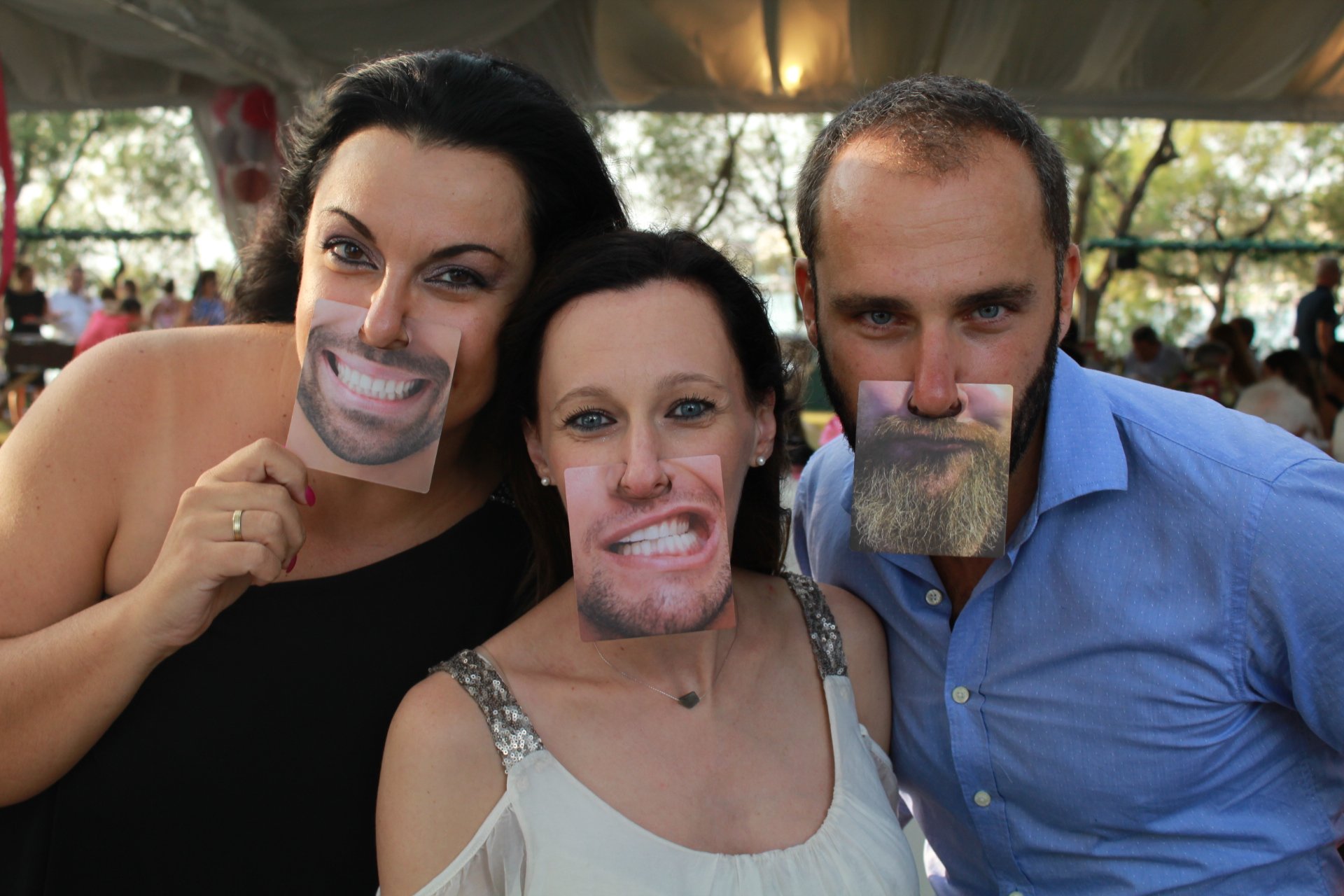Three people holding photo cards with exaggerated smiles and beards in front of their mouths during an outdoor event.